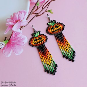Op de afbeelding: Een paar kralen pompoen oorbellen met een bungelend franje ontwerp. De oorbellen hebben oranje, gele, groene en zwarte kralen, met een jack-o'-lantern gezicht op de pompoen. De oorbellen staan tegen een roze achtergrond.