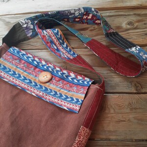 May include: A brown fabric tote bag with a patterned flap and strap. The flap features a blue, red, and orange geometric design, secured by a wooden button. The strap is a patchwork of floral and striped fabrics.