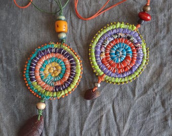 May include: Two circular pendants crafted from coiled fabric strips in vibrant shades of orange, purple, yellow, and green. Each pendant features small beads along the edge, a central spiral design, and a dangling dark brown seed-like charm.