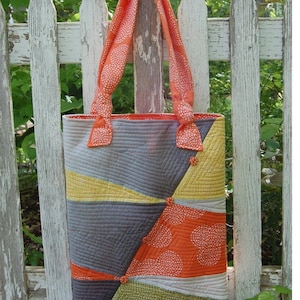 May include: A patchwork tote bag with a geometric design in shades of orange, yellow, gray, and green. The bag has a long strap and is made of fabric with a subtle pattern.