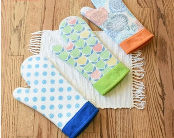 Op de afbeelding: Drie ovenwanten met kleurrijke patronen liggen op een witte mat met franjes. Eén want heeft blauwe stippen, een andere een groen en geel patroon en de derde een bloemenpatroon. De tekst "Hot Stuff Oven Mitt" is zichtbaar. De wanten zijn ongeveer 33 cm hoog en 19 cm breed.