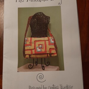May include: A colorful patchwork bag with geometric patterns in red, yellow, blue, and white. The bag is displayed on a mannequin and a stand. The text "The Amanda Bag" and "Designed by: Cynthia Tazelaar" is printed on the page.