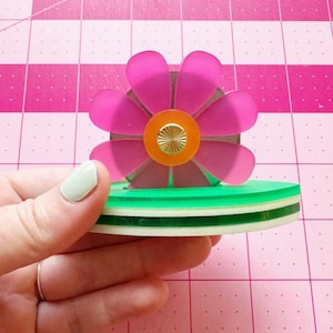 May include: A pink and green layered acrylic flower with a gold center. The flower is on a green and white layered base.