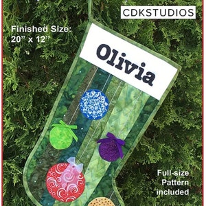 May include: A green and white Christmas stocking with a personalized name "Olivia" and colorful ornaments. The stocking is 20 inches by 12 inches and includes a full-size pattern.