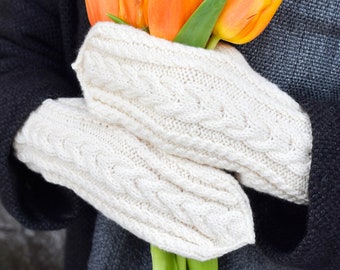 Könnte beinhalten: Cremefarbene Zopfmuster-Fäustlinge halten einen Strauß orangefarbener Tulpen. Die Fäustlinge sind aus weichem, strukturiertem Garn gefertigt. Die Person trägt einen dunkelgrauen Mantel und Schal. Die Tulpen haben grüne Stiele und Blätter.