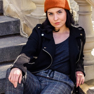 May include: A woman wearing an orange knitted beanie, a black jacket with silver zips, a grey v-neck shirt, and plaid trousers. She is sitting on stone steps with a white balustrade in the background.