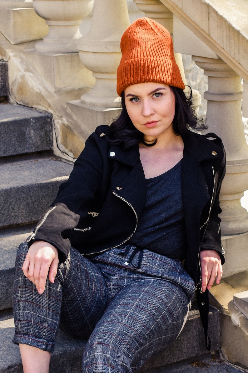 May include: A woman wearing an orange knitted beanie, a black jacket with silver zips, a grey v-neck shirt, and plaid trousers. She is sitting on stone steps with a white balustrade in the background.
