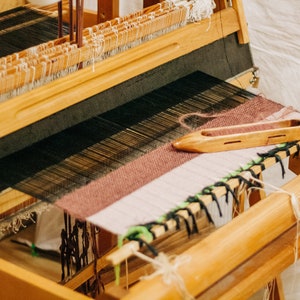 May include: A wooden loom with a partially woven fabric in shades of brown, white, and black. A wooden shuttle is resting on the fabric.