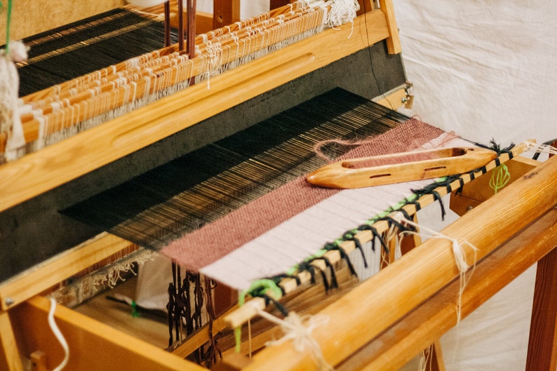May include: A wooden loom with a partially woven fabric in shades of brown, white, and black. A wooden shuttle is resting on the fabric.