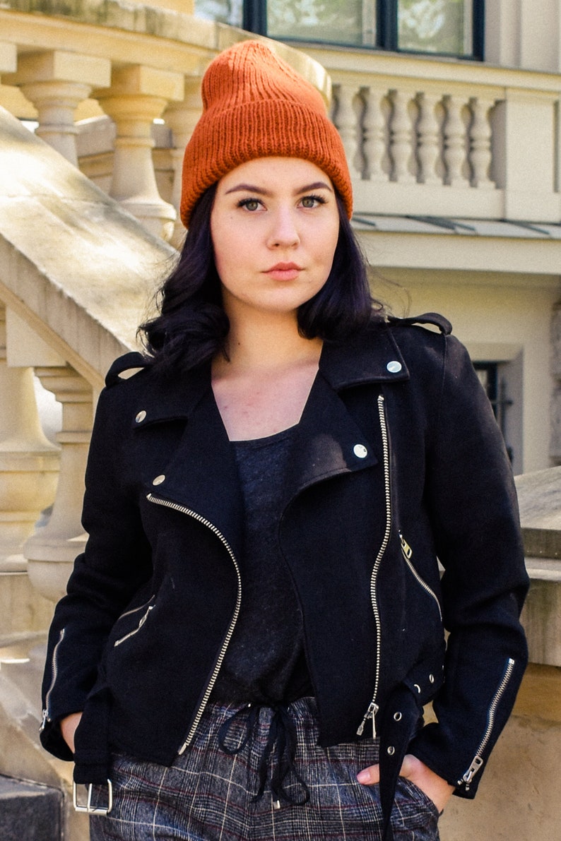 May include: A woman wearing a burnt orange knitted beanie, a black jacket with silver zips, and plaid trousers. The jacket has a classic biker style with multiple zips and a tailored fit. The outfit is completed with a dark grey top.