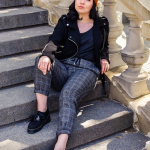 May include: A person wearing a black jacket, grey top, and plaid trousers sits on stone steps. They wear a burnt orange beanie and black shoes. The steps have a decorative stone railing.