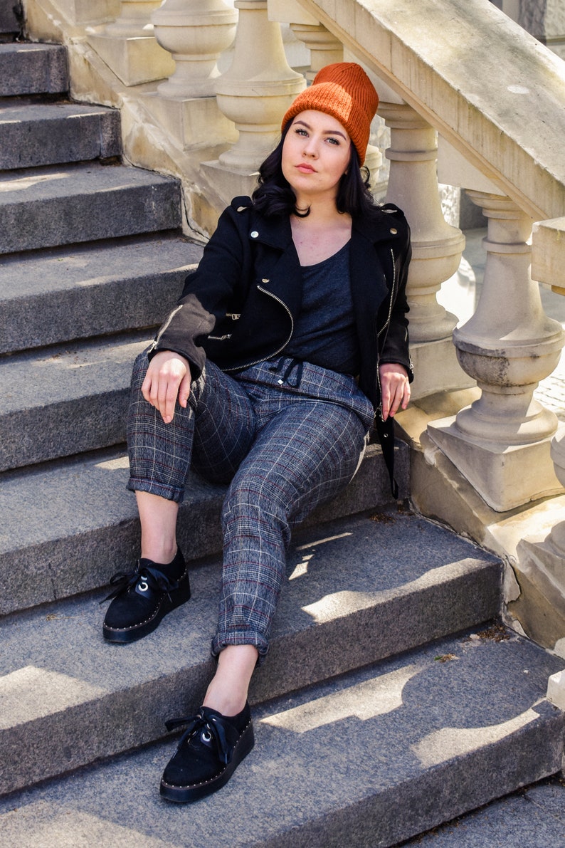 May include: A person wearing a black jacket, grey top, and plaid trousers sits on stone steps. They wear a burnt orange beanie and black shoes. The steps have a decorative stone railing.
