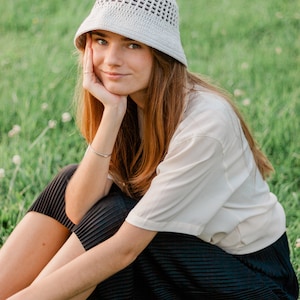 May include: A woman wearing a white crochet bucket hat sits on a grassy field. She is wearing a white t-shirt and a black pleated skirt.