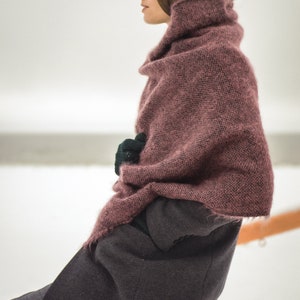 May include: A woman wearing a long, dark gray coat with a pink knitted scarf draped over her shoulders. She is sitting on a wooden railing with her hands tucked inside the scarf.