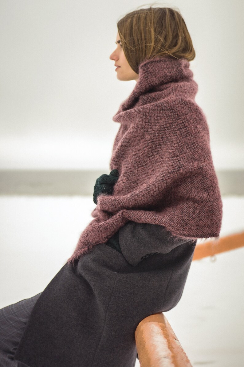 May include: A woman wearing a long, dark gray coat with a pink knitted scarf draped over her shoulders. She is sitting on a wooden railing with her hands tucked inside the scarf.