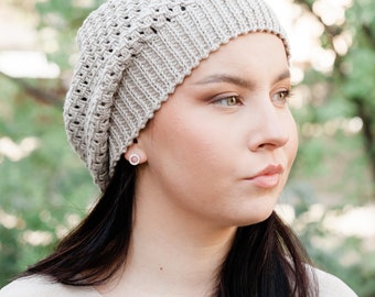 May include: A woman wearing a beige knitted beanie with a loose, open weave. The beanie is slouchy and has a relaxed fit. The woman has long dark hair and is looking off to the side.
