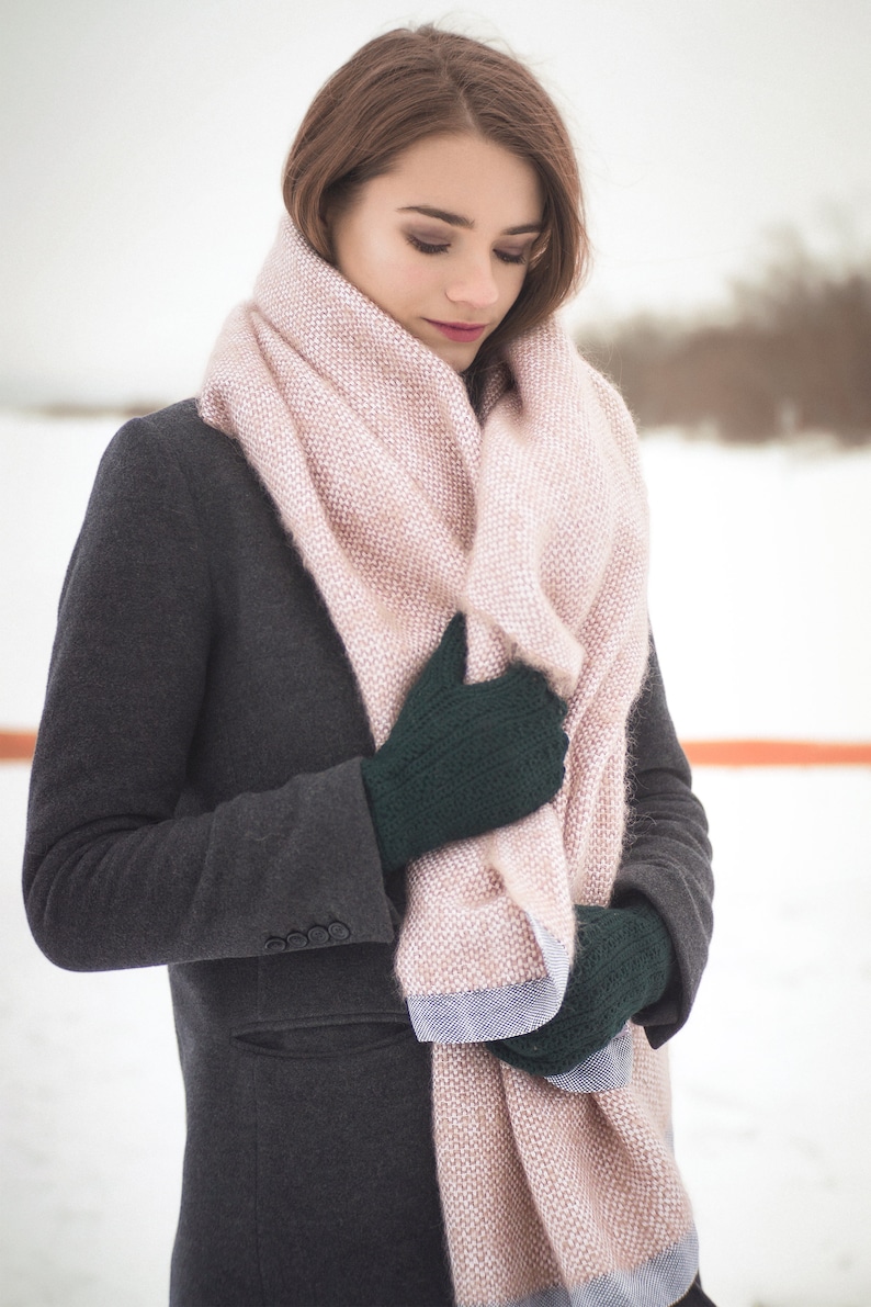 May include: A woman wearing a gray coat with a pink and white scarf wrapped around her neck. She is wearing dark green gloves and is holding the scarf in her hands.