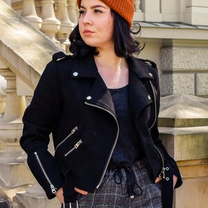 May include: A black biker jacket with silver zips and a burnt orange beanie. The jacket has an asymmetrical design with a notched collar and a belt. The trousers are plaid. The jacket is made of a wool blend.