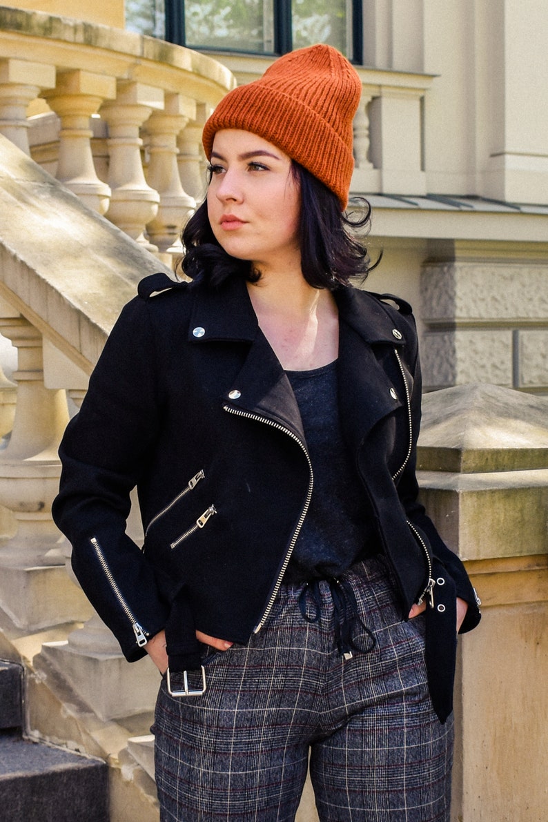 May include: A black biker jacket with silver zips and a burnt orange beanie. The jacket has an asymmetrical design with a notched collar and a belt. The trousers are plaid. The jacket is made of a wool blend.