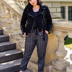 May include: A black jacket with silver zips, an orange beanie, and plaid trousers are worn. The outfit includes a dark grey top and black shoes. The background features a stone staircase and a building.