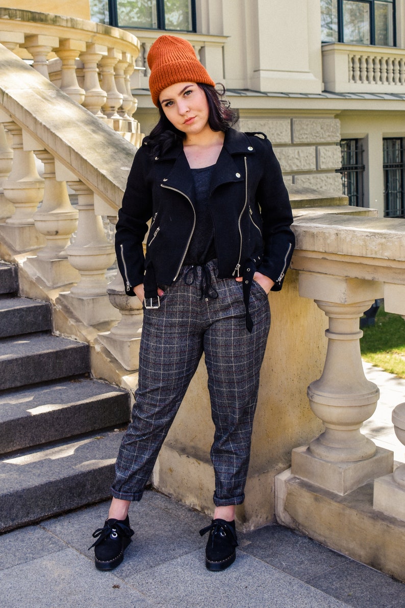May include: A black jacket with silver zips, an orange beanie, and plaid trousers are worn. The outfit includes a dark grey top and black shoes. The background features a stone staircase and a building.