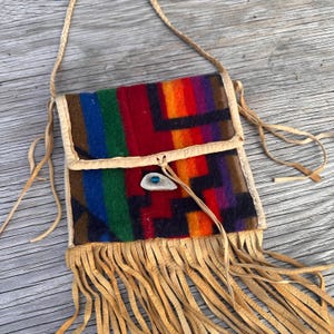 May include: A colorful, fringed crossbody bag with a geometric pattern in red, orange, purple, green, and blue. The bag has a leather strap and a decorative clasp with a blue stone. The fringe is a light tan color.