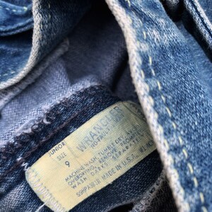 May include: A close-up of a blue denim jean label with white stitching. The label is white with black text that reads "Junior Size 9 Wrangler" and "Machine wash, tumble dry, avoid overdrying, remove promptly, wash darks separately, S9HPA381R Made in USA. WLAC".