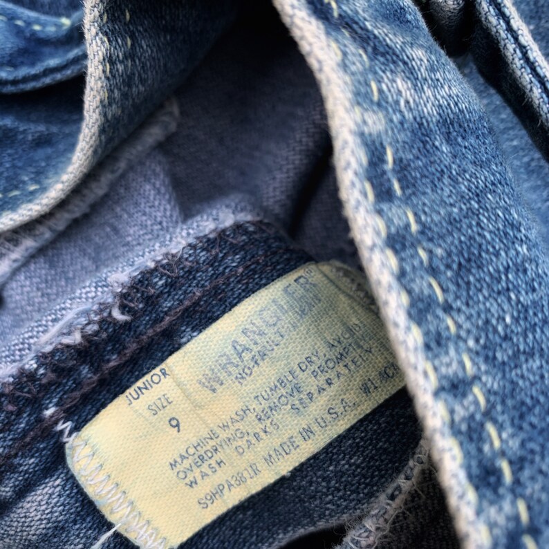 May include: A close-up of a blue denim jean label with white stitching. The label is white with black text that reads "Junior Size 9 Wrangler" and "Machine wash, tumble dry, avoid overdrying, remove promptly, wash darks separately, S9HPA381R Made in USA. WLAC".
