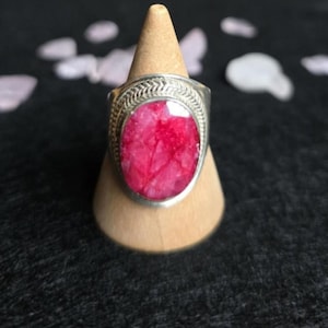 May include: A silver ring featuring a large, oval-cut, vibrant red gemstone. The ring has a detailed, patterned border. It is displayed on a small, wooden cone-shaped stand against a dark backdrop.