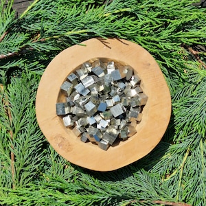 Peut inclure: Un bol en bois rempli de petits cristaux de pyrite cubiques, brillants et métalliques. Le bol est entouré de feuillage vert.