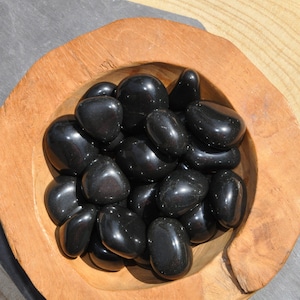 Peut inclure: Un bol en bois rempli de pierres lisses en obsidienne noire roulées. Les pierres sont polies et ont une finition brillante.