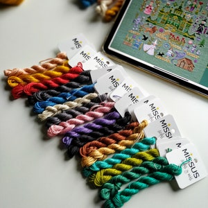 May include: A collection of embroidery floss skeins in various colours, including red, orange, yellow, green, blue, purple, and brown. The skeins are arranged in a row, with the labels "Miss Miss" and "Missus" visible.