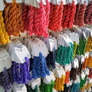 May include: A display of colorful embroidery floss in various shades, including yellow, orange, pink, red, blue, green, and purple. Each skein is attached to a white tag with a number. The floss is arranged on a pegboard.