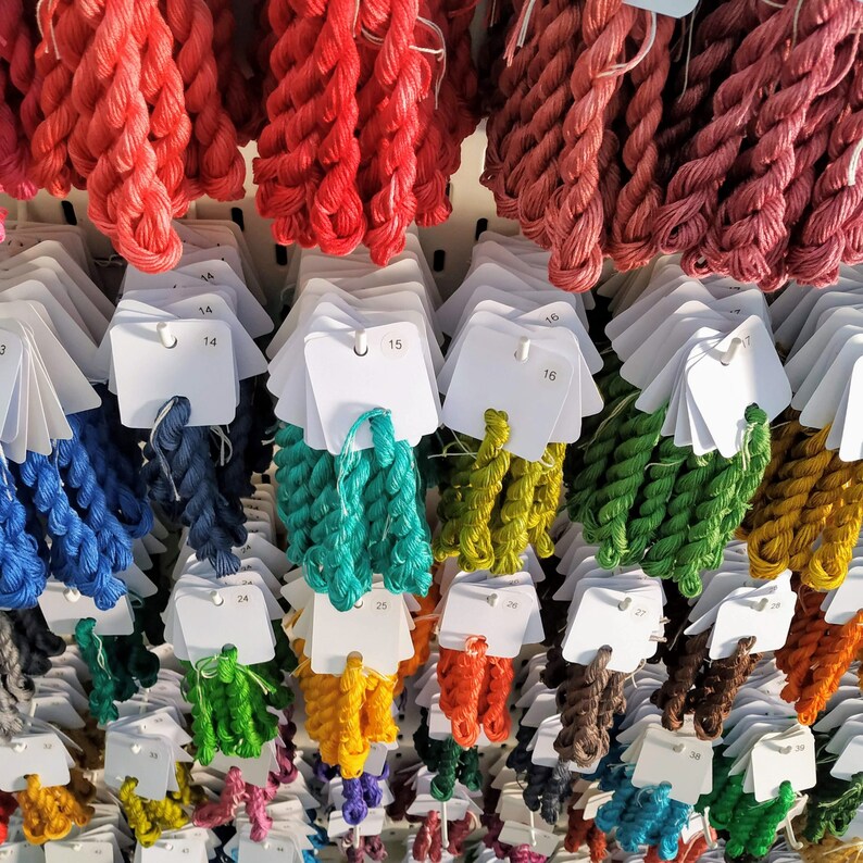 May include: A display of colorful embroidery floss in various shades, including red, blue, green, and yellow. Each skein is attached to a white tag with a number. The floss is arranged in neat rows, creating a visually appealing and organized presentation.