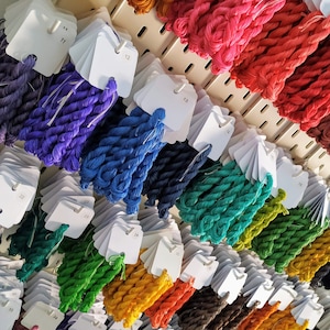 May include: A display of colorful embroidery floss in various shades, including purple, blue, green, yellow, orange, and red. Each color is neatly organized with a white tag, creating a visually appealing arrangement for craft enthusiasts.