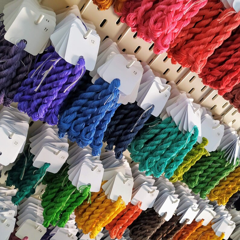 May include: A display of colorful embroidery floss in various shades, including purple, blue, green, yellow, orange, and red. Each color is neatly organized with a white tag, creating a visually appealing arrangement for craft enthusiasts.