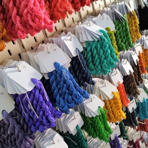 May include: A display of colorful embroidery floss in various shades, including pink, red, blue, green, and yellow. Each skein is attached to a white tag with a number. The floss is arranged on a pegboard, creating a visually appealing arrangement.