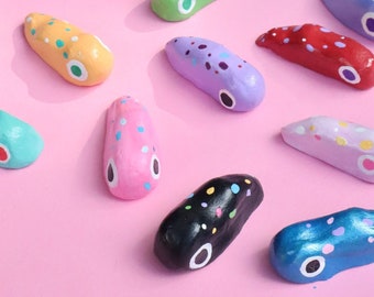 Puede incluir: Una colección de objetos decorativos oblongos pintados a mano, con diseños de puntos y ojos. Los objetos son de varios colores, incluyendo rojo, azul, verde, amarillo y morado, y están dispuestos sobre un fondo rosa. Medidas no disponibles.