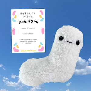 Pode incluir: Uma criatura de pelúcia branca e texturizada com dois olhos de contas pretas e uma pequena boca costurada, acompanhada de um cartão branco que diz: thank you for adopting BING BONG, - scared of fireworks, - loves cartoons, - one soft serve ice cream away from spiraling into madness, GOBLIN PUPPY STUDIOS.