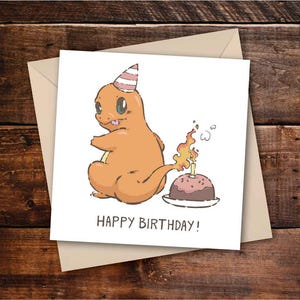 May include: A birthday card featuring a cartoon Charmander character wearing a party hat, with a lit birthday cake. The card reads "HAPPY BIRTHDAY!" in black text. The card is on a beige envelope, set against a wooden background.