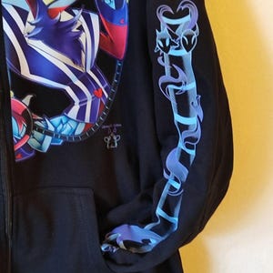 May include: Black zippered hoodie with a blue and purple graphic print of a cartoon character on the front and a blue and purple graphic print of a stylized heart on the sleeve.