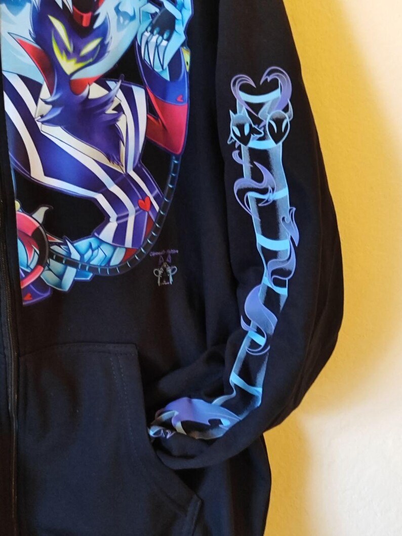 May include: Black zippered hoodie with a blue and purple graphic print of a cartoon character on the front and a blue and purple graphic print of a stylized heart on the sleeve.