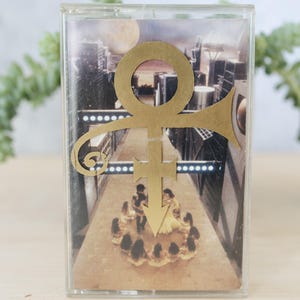 May include: A vintage cassette tape featuring a gold ankh-like symbol with a trumpet. The cassette has a cityscape background with figures in the foreground. The tape is in a clear plastic case, resting on a light wooden surface.