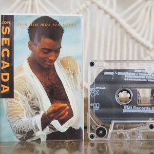 May include: A clear plastic cassette tape case with a black cassette tape inside. The cassette tape has the text "JON SECADA" and "Otro Dia Mas Sin Verte" printed on it. The cassette tape is labeled "EMI Records Group".