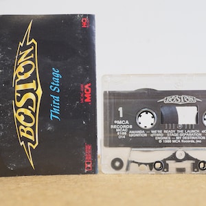 May include: A cassette tape with a black and white label that says "BOSTON" and "Third Stage". The cassette tape is in a clear plastic case. The label also includes the text "MCA Records", "MCAC", "Amanda", "We're Ready The Launch", "Countdown", "Third Stage Separation", "Engines - My Destination", "Cool The", and "1986 MCA Records, Inc."