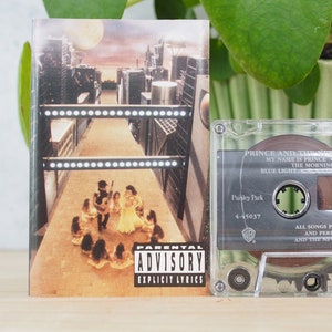 May include: A cassette tape of Prince and The New Power Generation's album "Paisley Park". The album cover features a group of people in a circle with a city skyline in the background. The cassette tape is clear plastic with a black label. The label includes the album title, artist, and track list. The label also includes a parental advisory warning.