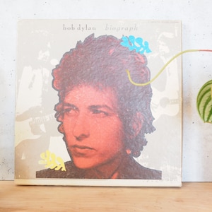 May include: A vintage vinyl record album cover featuring a close-up portrait of Bob Dylan. The album title "Biograph" is printed in white letters on a grey background. The cover features colourful abstract shapes.
