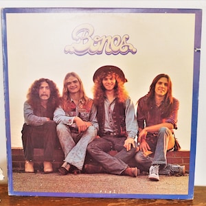 May include: A vintage vinyl record album cover featuring a band called "Bones". The cover art shows four men sitting on a brick wall, with a sunset in the background. The band members are all wearing casual clothing, including jeans and denim jackets.