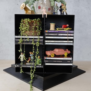 May include: A black, multi-compartment shelf unit displays various items. It features a small pink toy car, miniature figurines, cassette tapes, and a small gold gramophone. A trailing plant adds a touch of greenery.