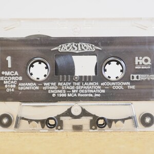 May include: A clear plastic cassette tape case with a black cassette tape inside. The cassette tape has the text "BOSTON" in a silver font on the top. The cassette tape has the text "MCA RECORDS MCAC 6188 014" on the left side and the text "AMANDA - WE'RE READY THE LAUNCH: a) COUNTDOWN IGNITION c) THIRD STAGE SEPARATION - COOL THE ENGINES - MY DESTINATION &copy; 1986 MCA Records, Inc" on the right side. The cassette tape also has the text "HQ HIGH QUALITY" on the right side.